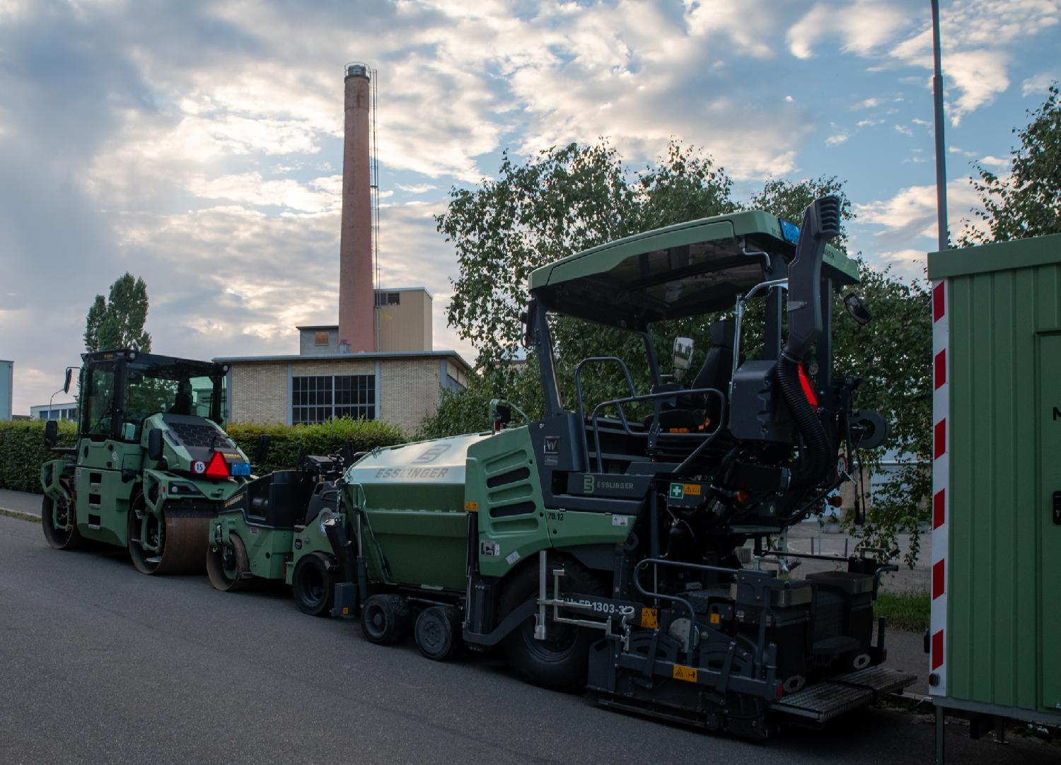 Some light-green construction vehicles in front of an industrial building with a high brick chimney.