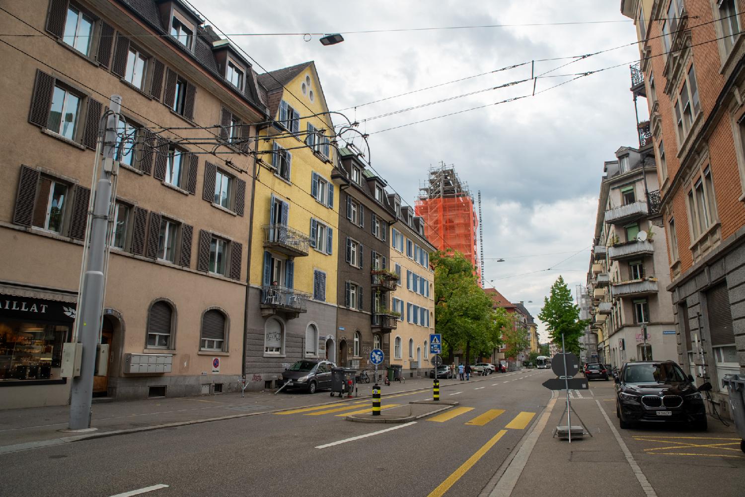 A two-way street with 4-storey buildings and a church with bright orange scaffolding in the background.