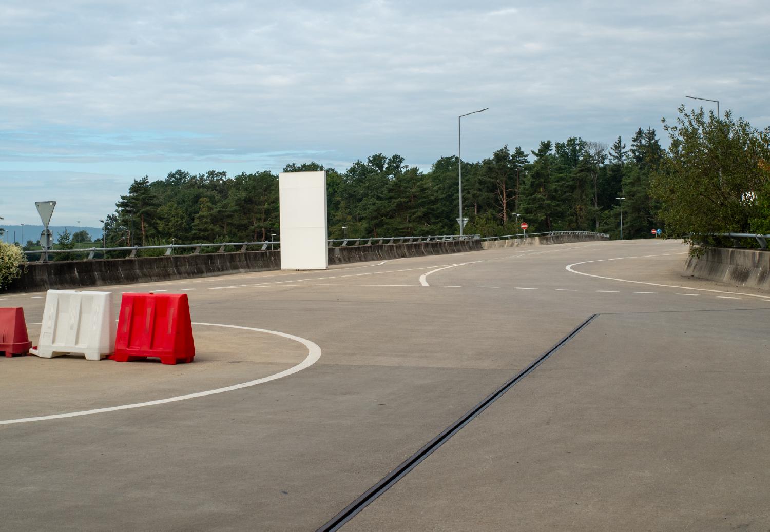 A car road street ending on a roundabout with white and red works blocks. There is trees behind the road.