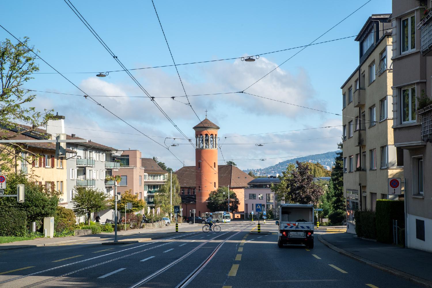 A two-way street with 3-4-storey buildings on each side, tram tracks in the middle, and a brick-red church tower in the background.