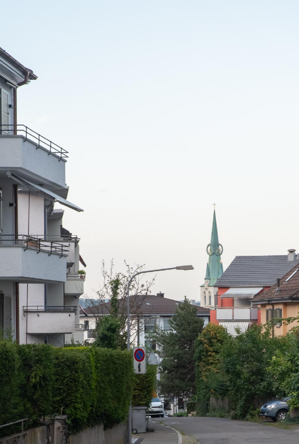 Allenmoosstrasse in Zürich: a street going downwards with 3-4 story buildings on each side, and the green steeple of the Herz Jesu (Zürich-Oerlikon) with its large clock faces in the background.