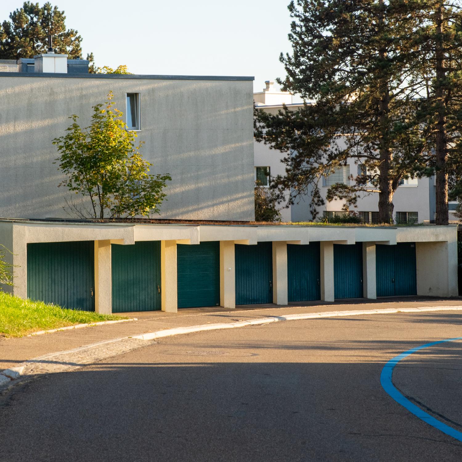 7 identical garage doors, painted in blue, in the curve of a road with a blue line. There are 3-story residential buildings and pine trees behind the garages.