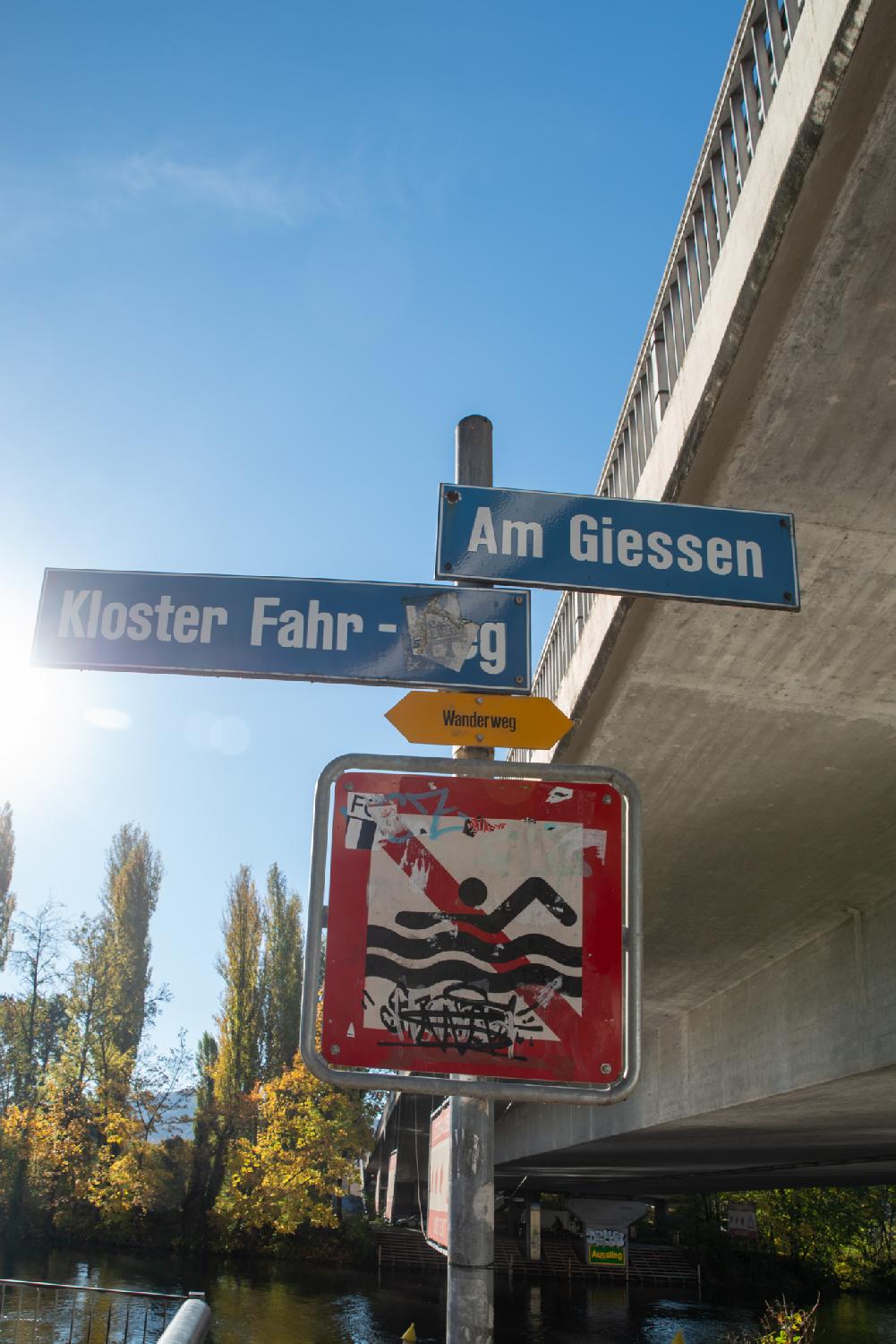 A grey pole with signs for Am Giessen and Kloster Fahr-Weg, a Wanderweg yellow sign, and a sign to indicate that it is forbidden to swim in the river.