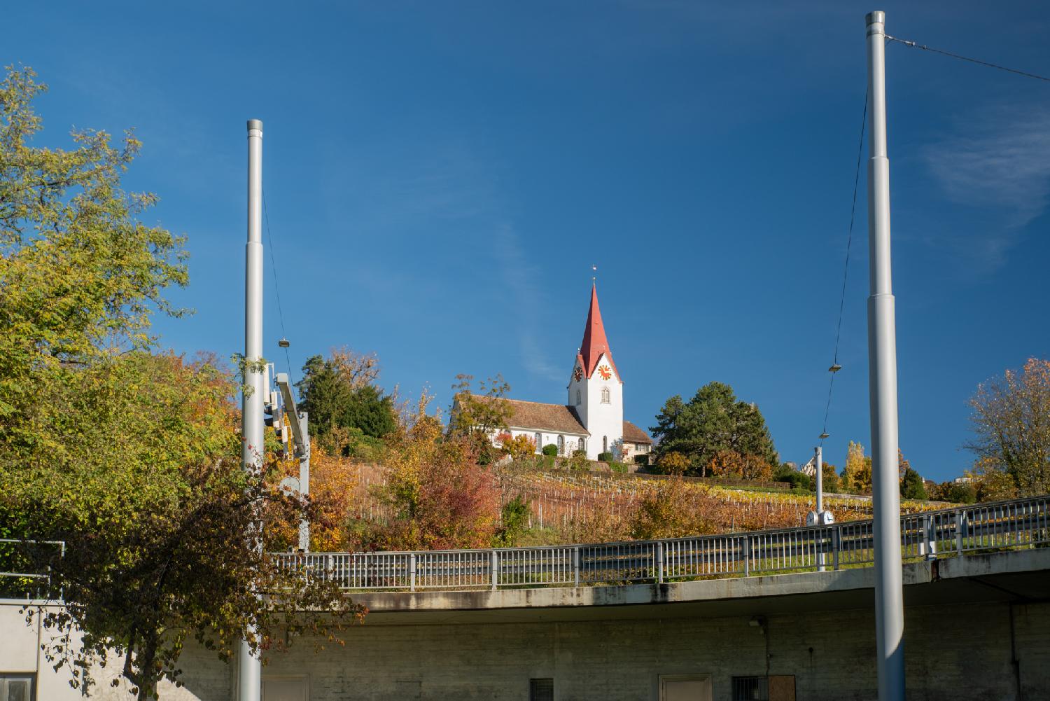 Seen from a low point: the top of a bridge with a metallic fence, vineyard in orange/red automn colors, and a white church with a red roof