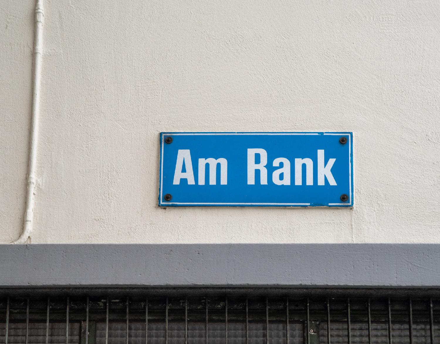 Blue "Am Rank" sign on a white wall with a lower grey border and a black grid below it