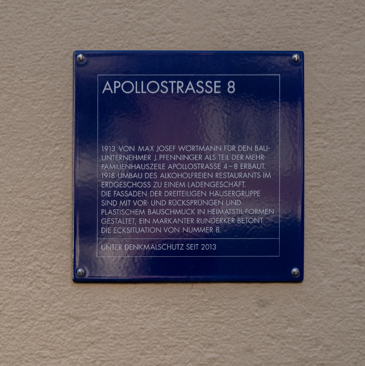 A blue sign that reads "1913 von Max Josef Wörtmann für den Bauunternehmer J. Pfenninger als Teil des Mehrfamilienhauszeile Apollostrasse 4-8 erbaut. 1918 Umbau des alkoholfreien Restaurants im Erdgeschoss zo einem Ladengeschäft. Die fassaden der dreiteiligen Häsergruppe sind mit Vor- und Rücksprüngen und plastischem Bauschmuck in Heimatstil-Formen gestaltet, ein markenter Runderker betont die Ecksituation von Nummer 8. Unter Denkmalschutz seit 2013."
