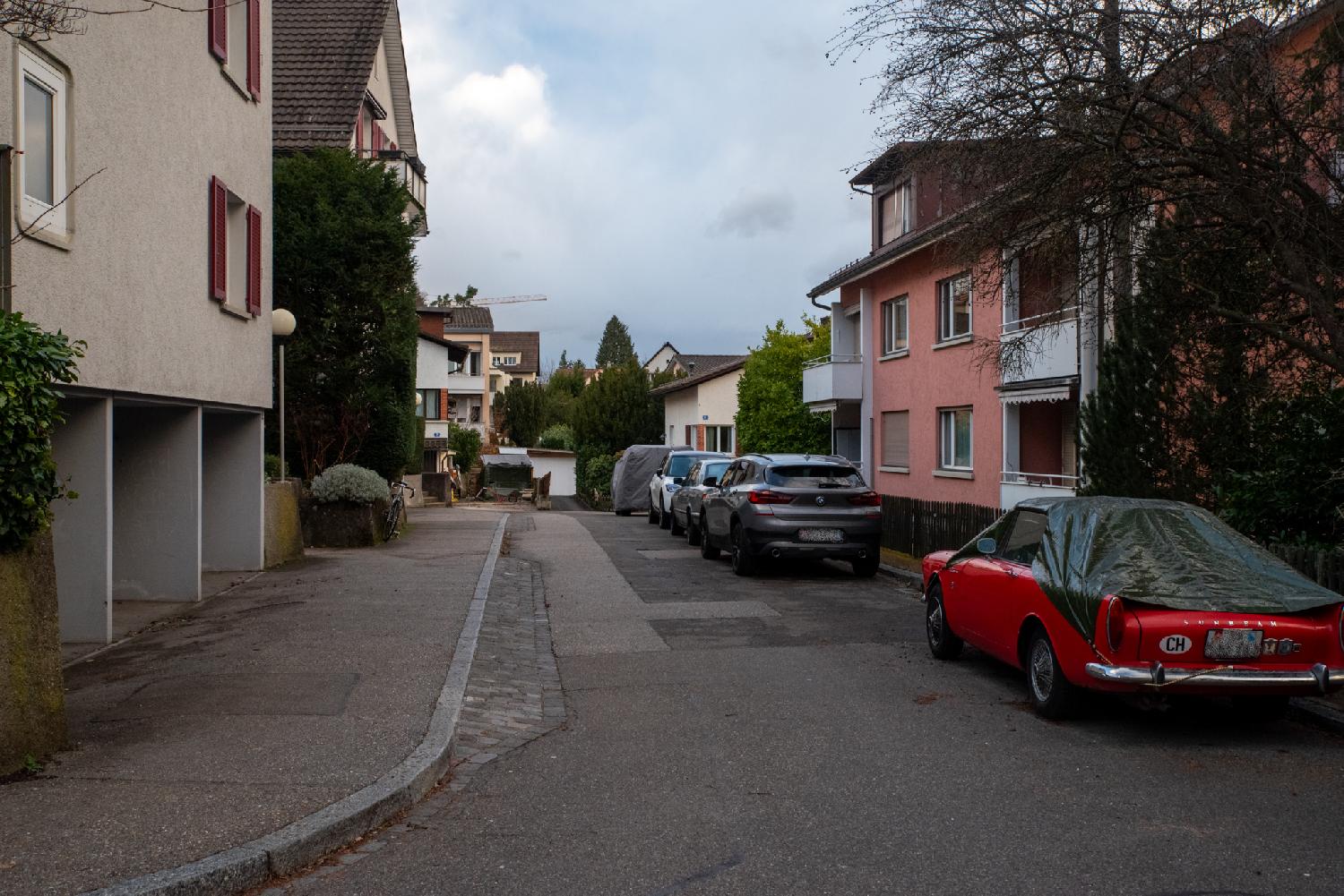 A residential street with three-story buildings and a row of cars on the right side