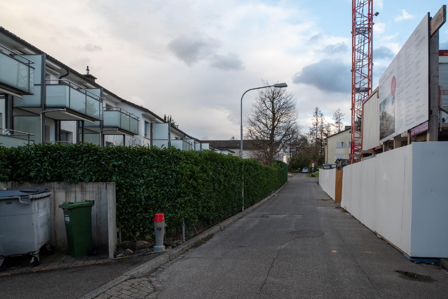 On the left, 2-story buildings with glass balconies behind a hedge. On the right, a construction site behind a white construction fence.
