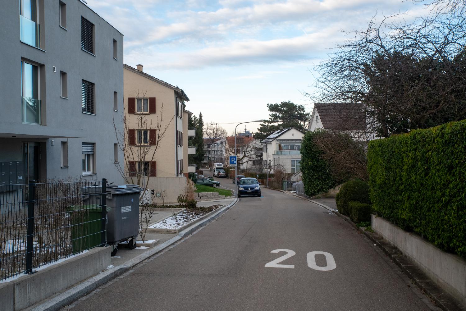 A narrow street with a 20km/h speed limit. There are 3-4-story buildings on both sides; the right side additionally has green hedges.
