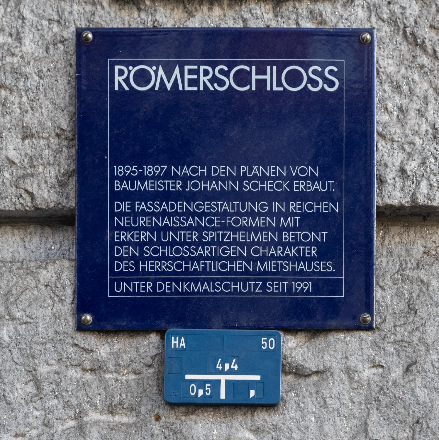 Blue sign written in German, reading "Römerschloss - 1895-1897 nach den Plänen von Baumeister Johan Scheck erbaut.  Die Fassadengestaltung in reichen Neurenaissance-Formen mit Erkern unter Spitzhelmen betont den schlossartigen Charakter des herrschaftlichen Mietshauses. Unter Denkmalschutz seit 1991."