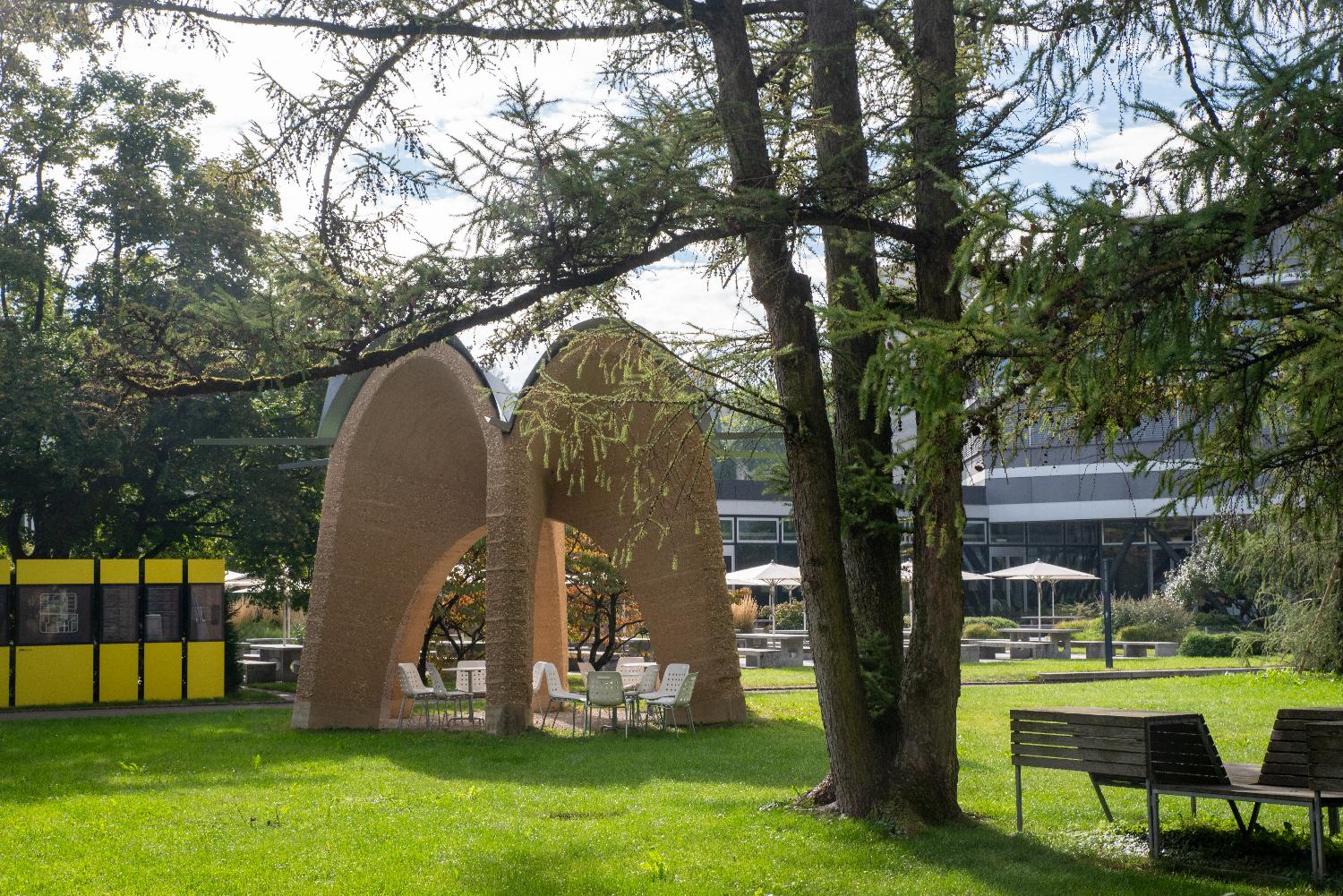 A lawn between paths, with a few sittings areas - one under sunshades, one under a stone structure, and one under trees.