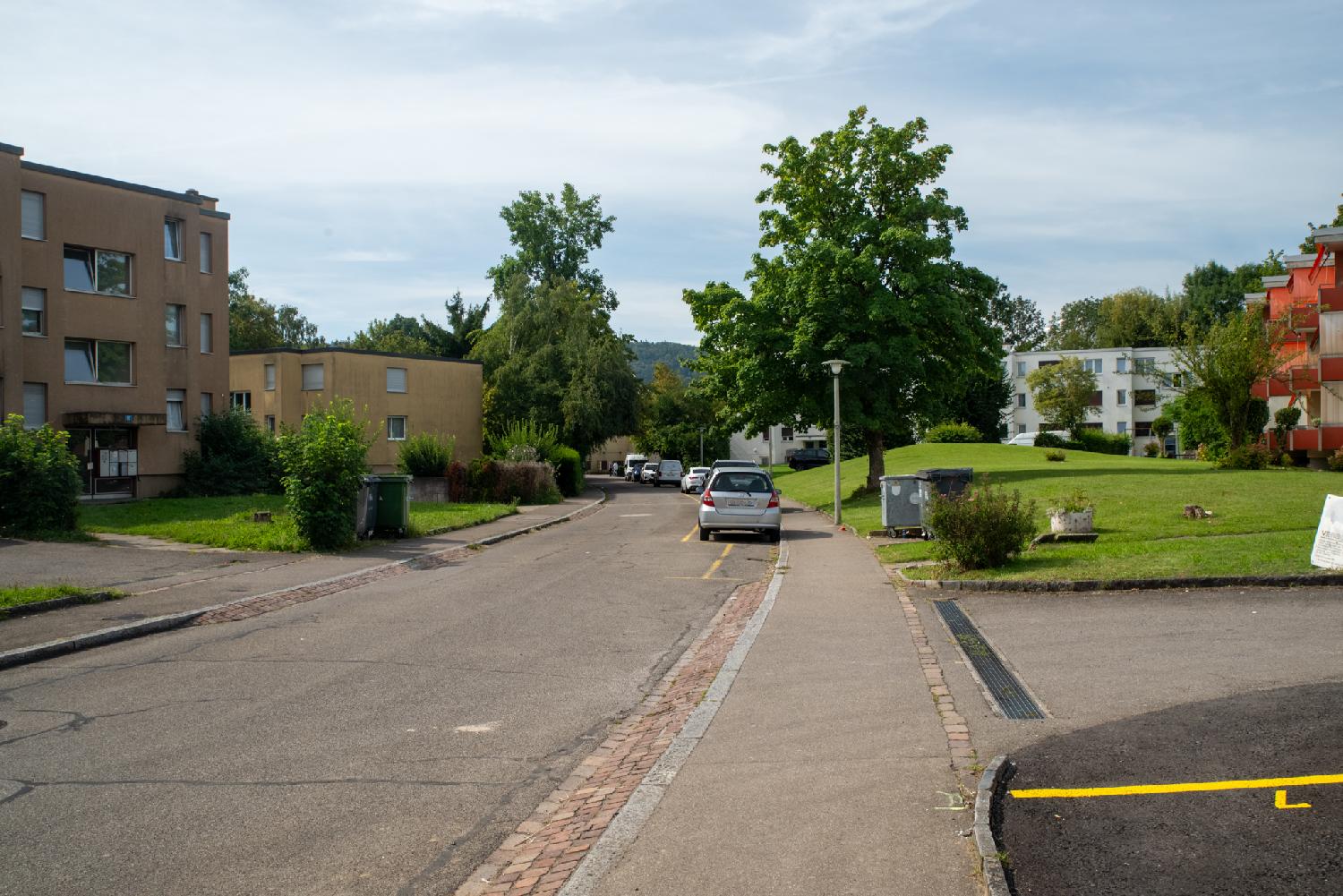 A residential street with 3-4-storey buildings, some trees and a green lawn. A row of cars is parked on the right side.