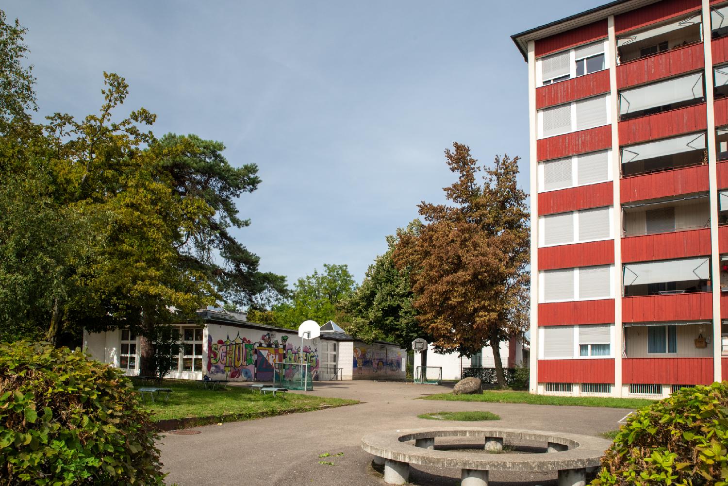 A small square with a round bench in front of a school building with painted walls and a 6-story residential building with red balconies