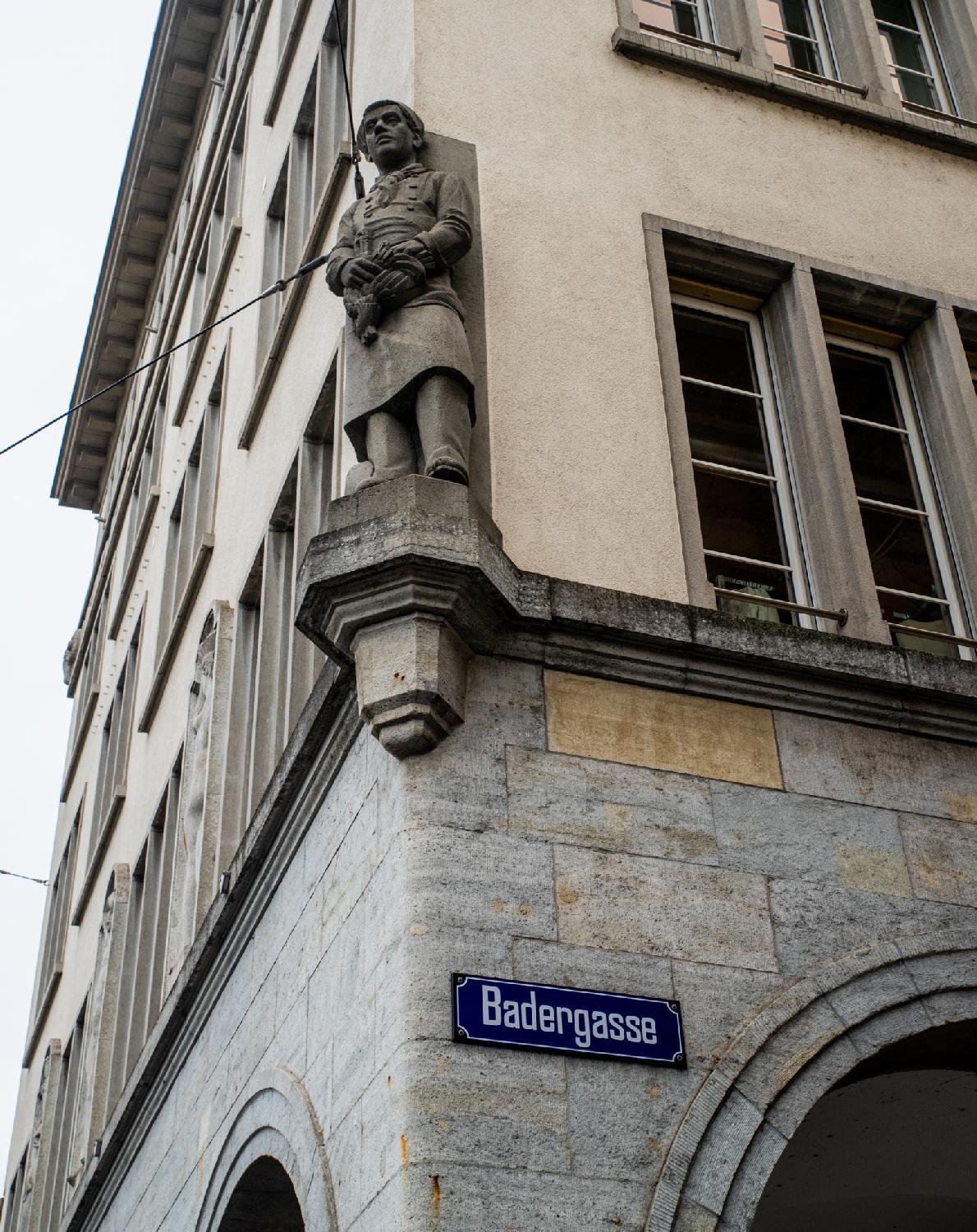 The corner of a building, with the statue of a work uniform man holding (maybe) a chicken. A blue "Badergasse" street name plate is attached to the building next to an arch.