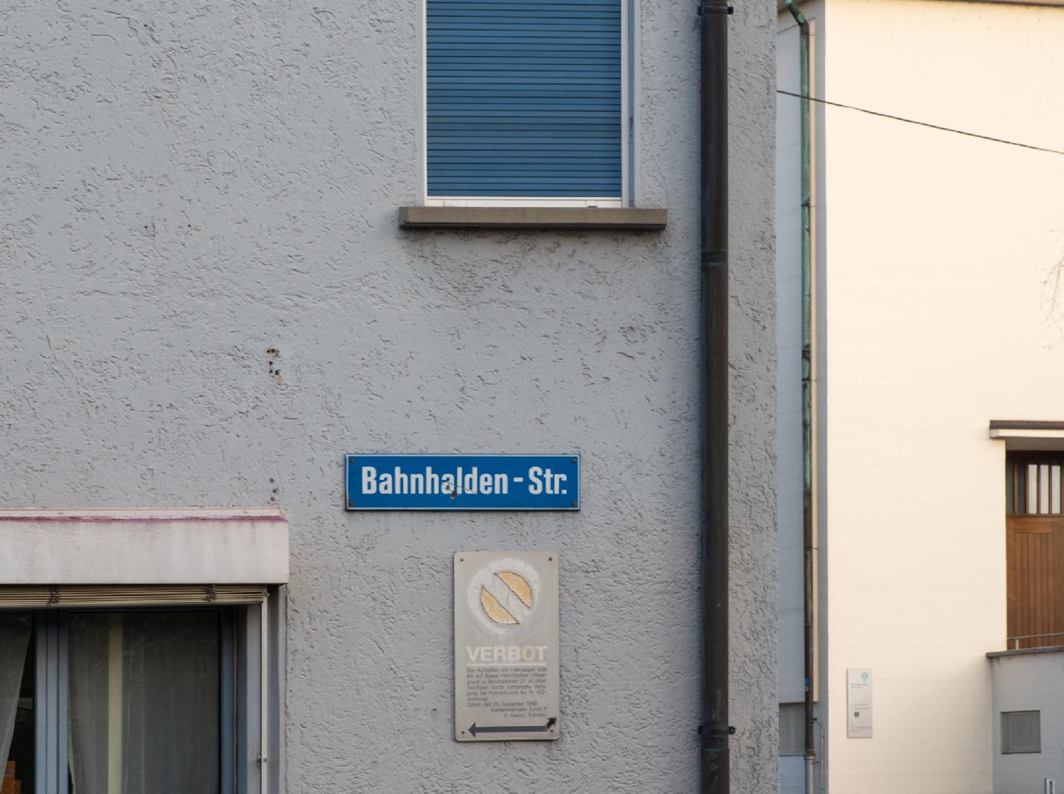 Blue street sign for Banhaldenstrasse, fixed to a grey-white wall of a building, with a "no parking" sign below it.