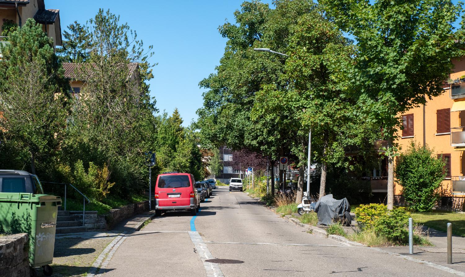 A narrow street with a row of cars parked on the left. There are 4-story buildings on each side of the road, hidden behind green trees.