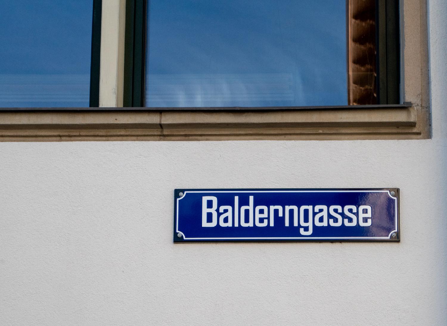 A blue Balderngasse street sign on a beige wall below a window.